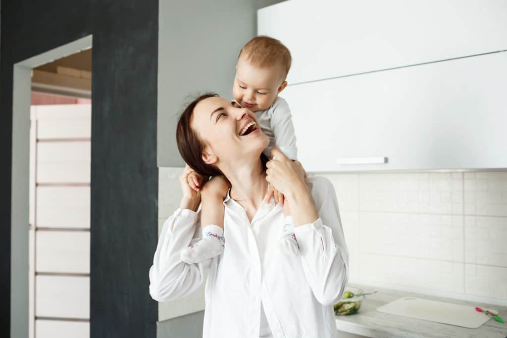 Portrait of happy young mother holding her little precious child on shoulders, looking at him and laughing while baby smiling - Learning to go with the flow