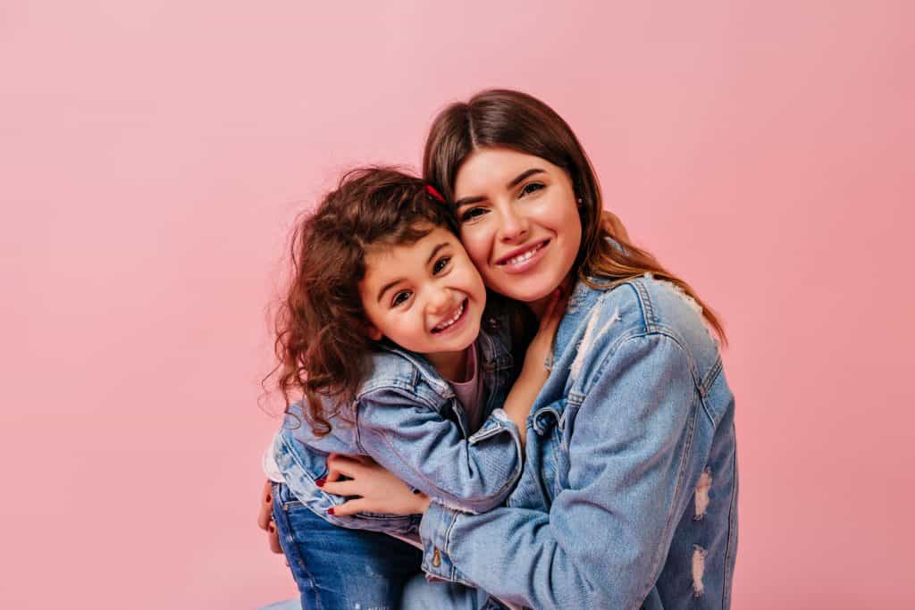 Emotional Self Care - Laughing mother and daughter looking at camera. Front view of young woman with preteen child isolated on pink background