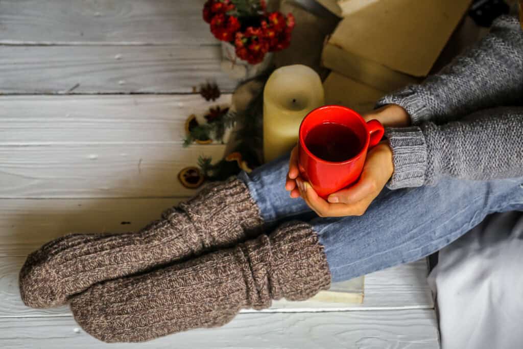 Warm and cozy,and warm concept girls feet in warm knitted socks with a Cup of tea over white wooden table with book and candle - Setting intentional goals for the new year